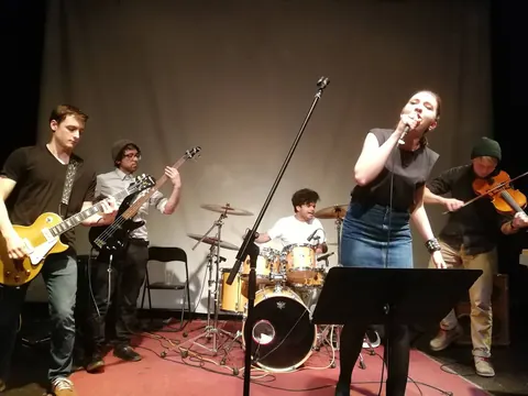 The band mid-song on a small stage — two guitars, a fiddle and a singer